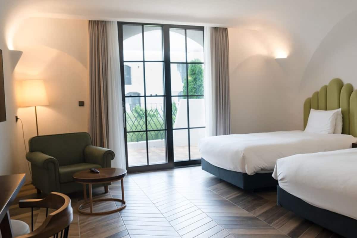 A hotel room with two beds, a sofa, a small table, and a large window with curtains.