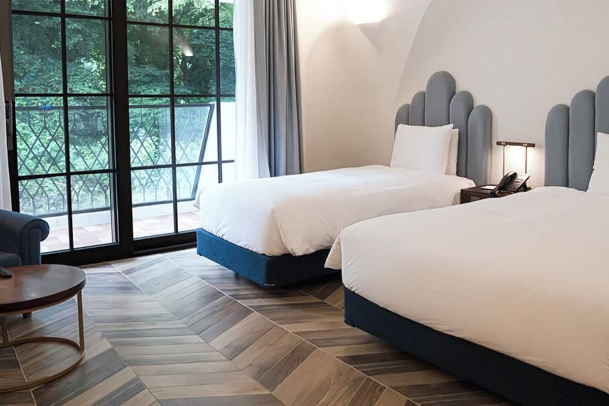 A hotel room with two beds, a sofa, a small table, and a large window with a view of greenery.