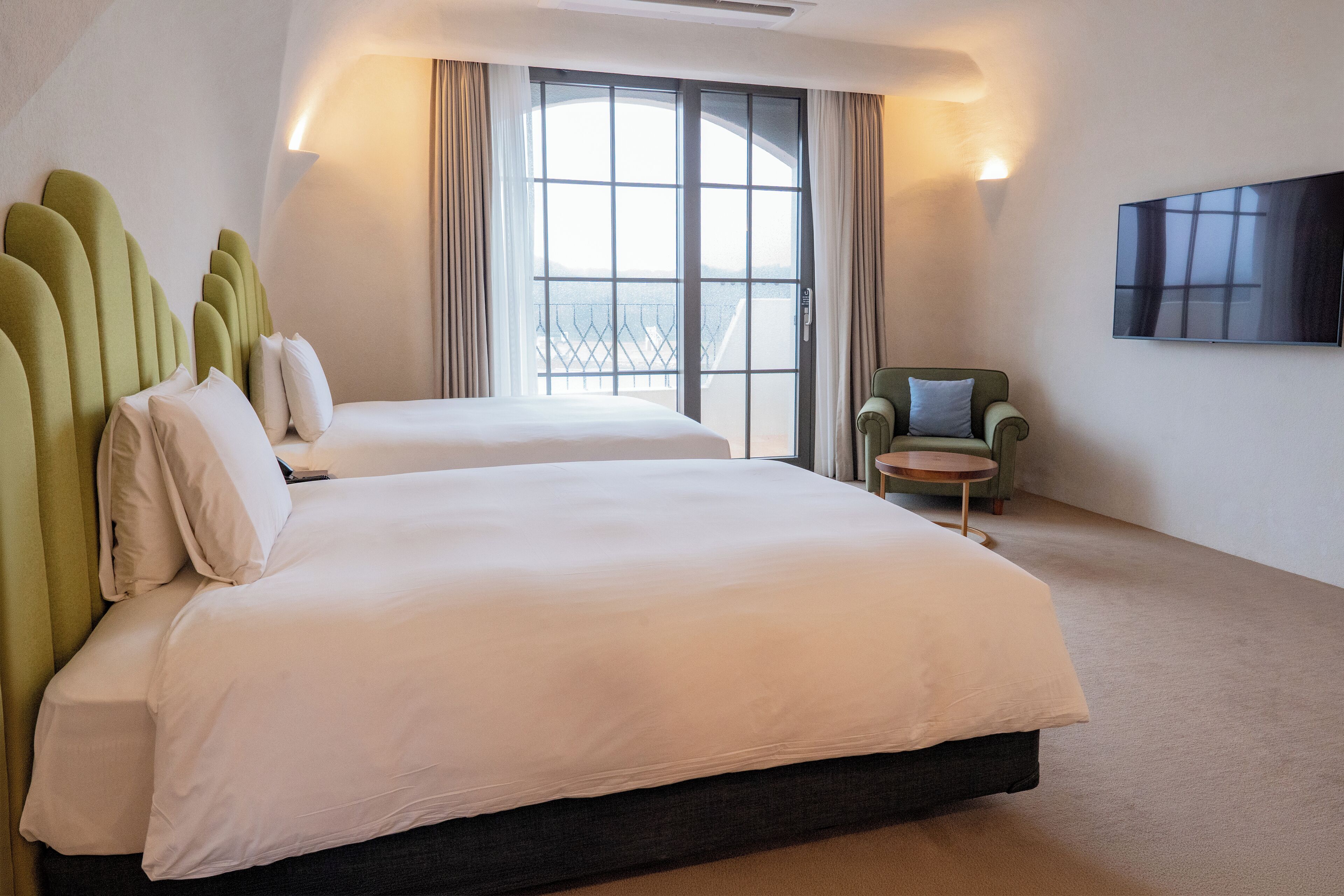 A hotel room with two beds, a TV, a chair, and a balcony with a view.