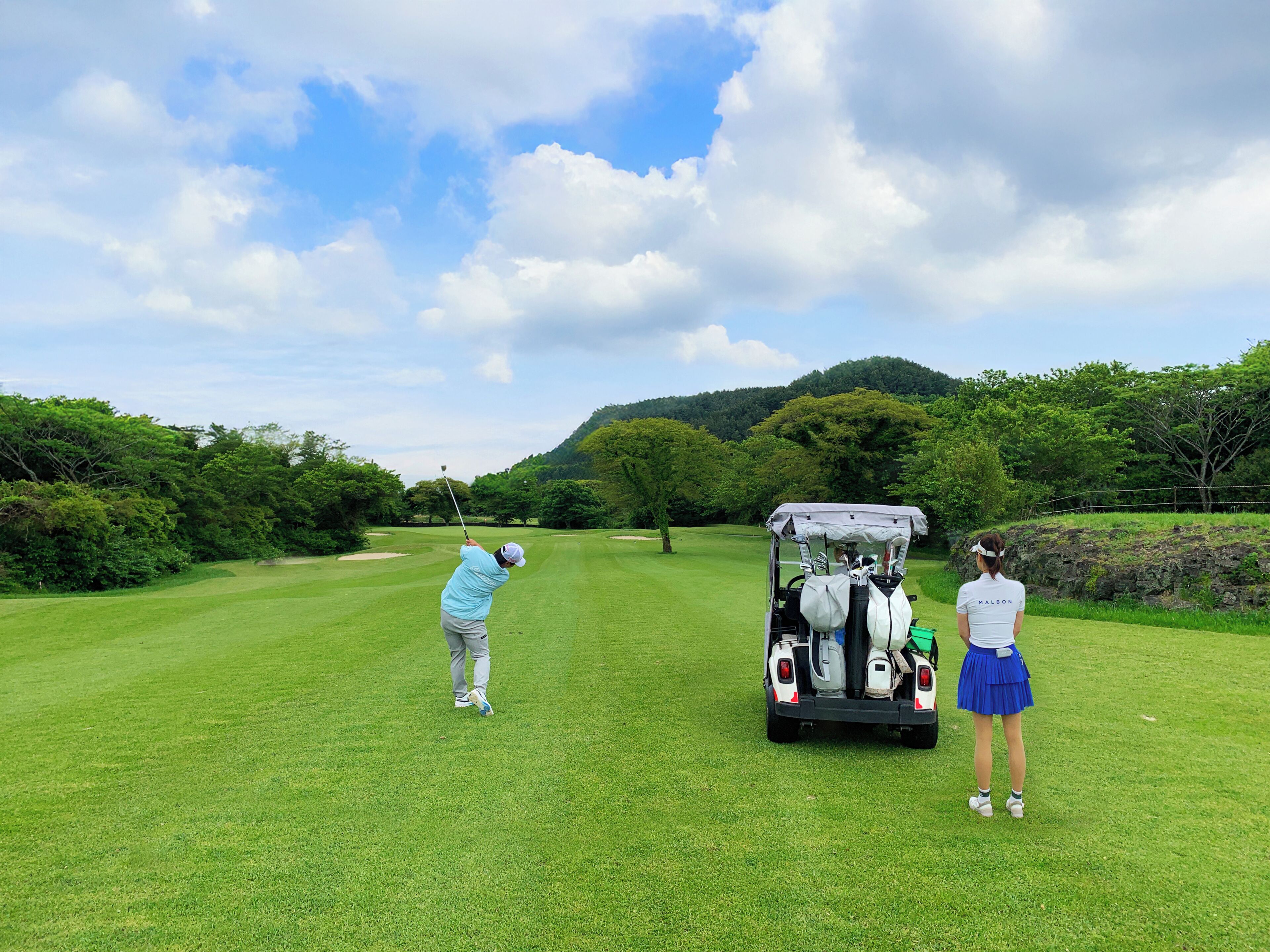 A golf course with a cart and two individuals, one preparing to hit a golf ball.