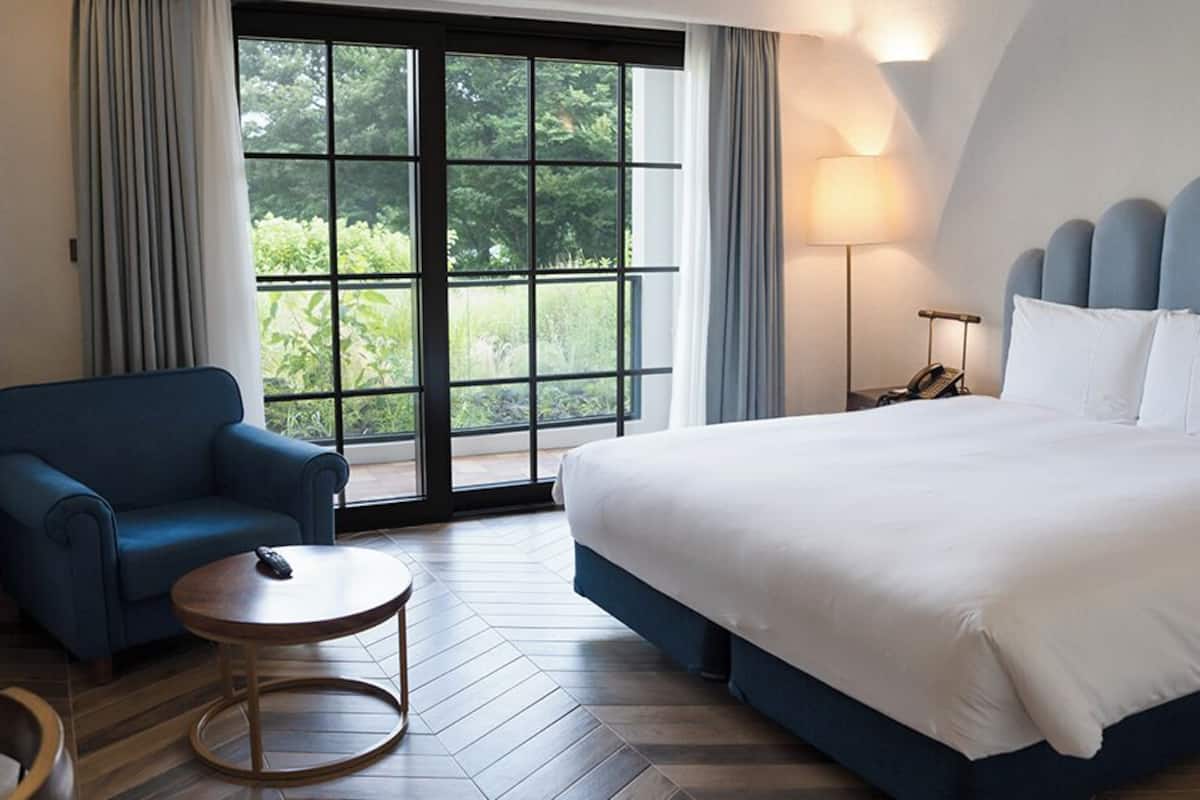 A hotel room with a large bed, a blue armchair, a small round table, and a large window offering a view of greenery.