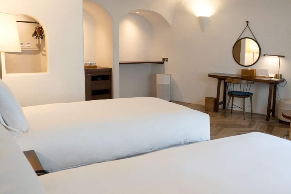 A hotel room with two beds, a desk with a chair, a small table, and a TV mounted on the wall.