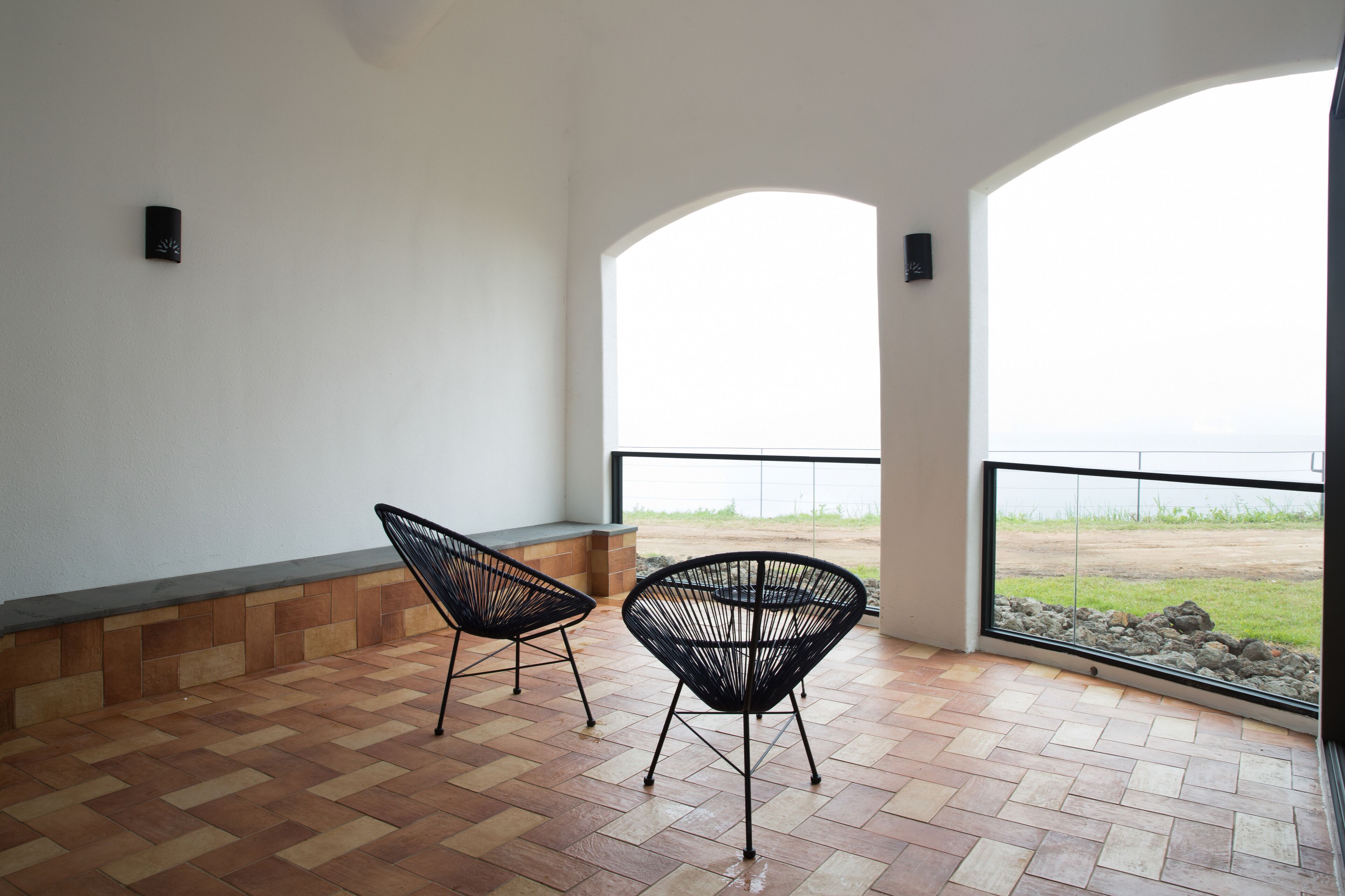 Un balcon avec deux chaises noires, un sol carrelé et une vue sur la plage, visible à travers de grandes fenêtres.