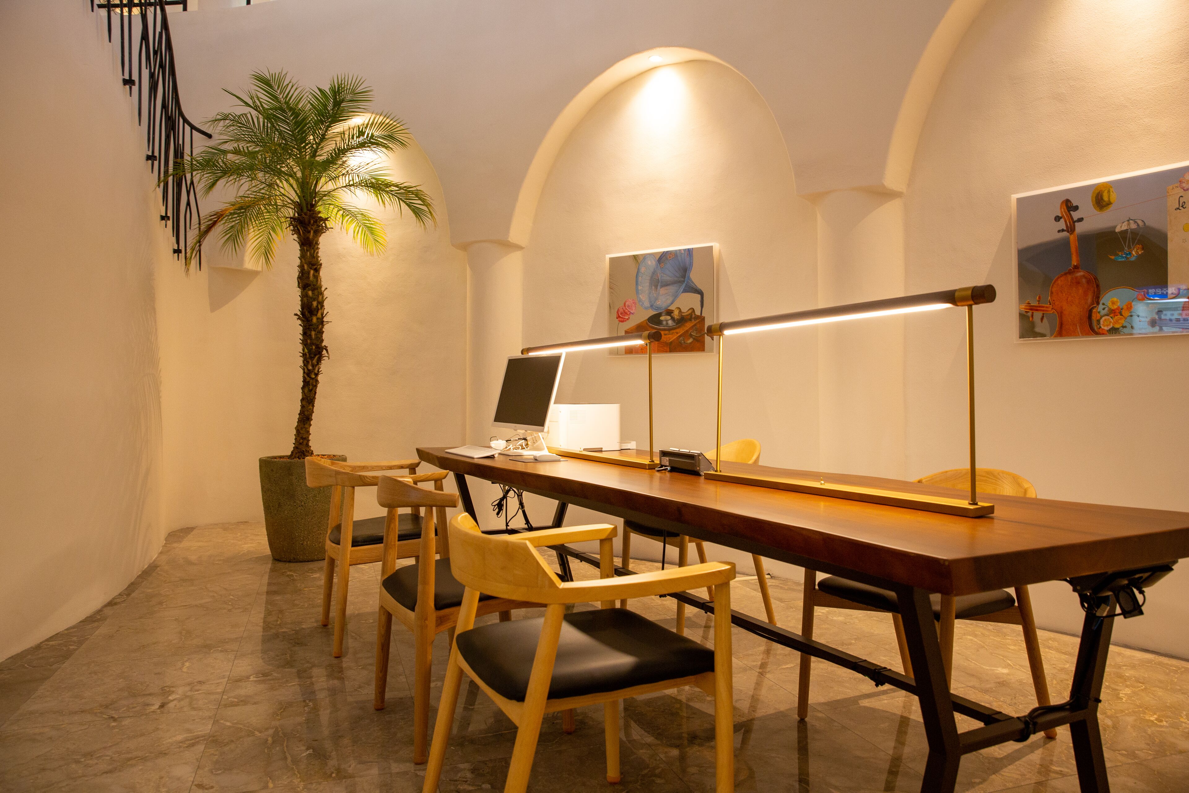 A conference room with a long table, four chairs, a laptop, a desk lamp, a potted plant, and a painting on the wall.