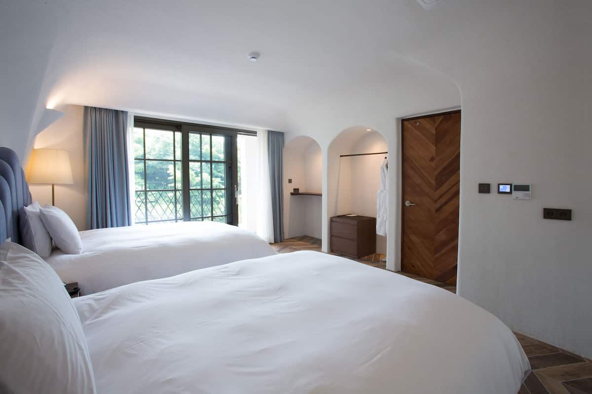 A hotel room with two beds, a large window with curtains, a wooden herringbone door, and a small dresser.