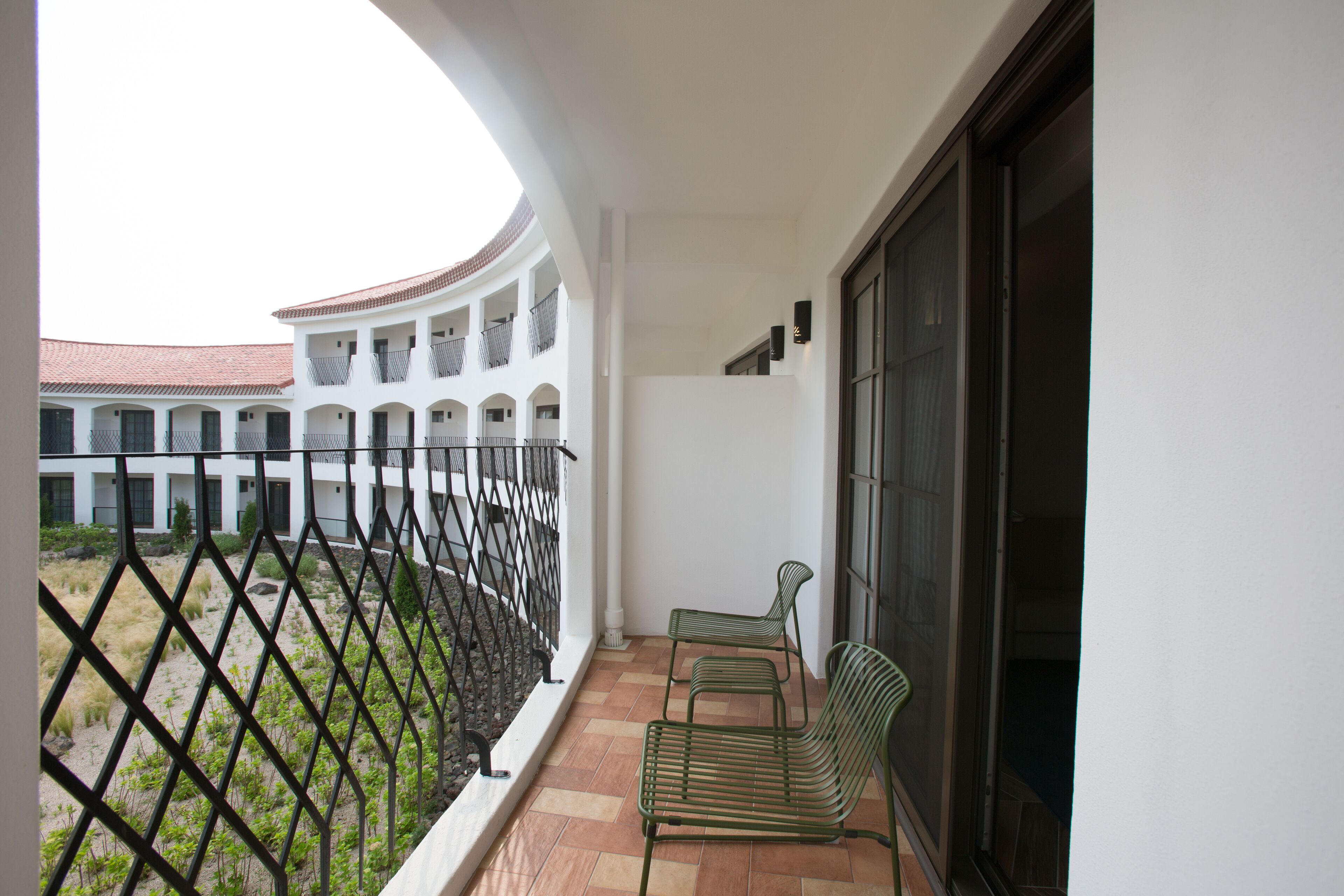 A balcony with two green metal chairs, a white wall, and a view of a courtyard with a building in the background.