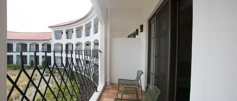 A balcony with two green metal chairs, a white wall, and a view of a courtyard with a building in the background.