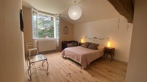 Un dormitorio con cama, lámparas de noche, una silla, un radiador y una ventana con vistas a árboles.