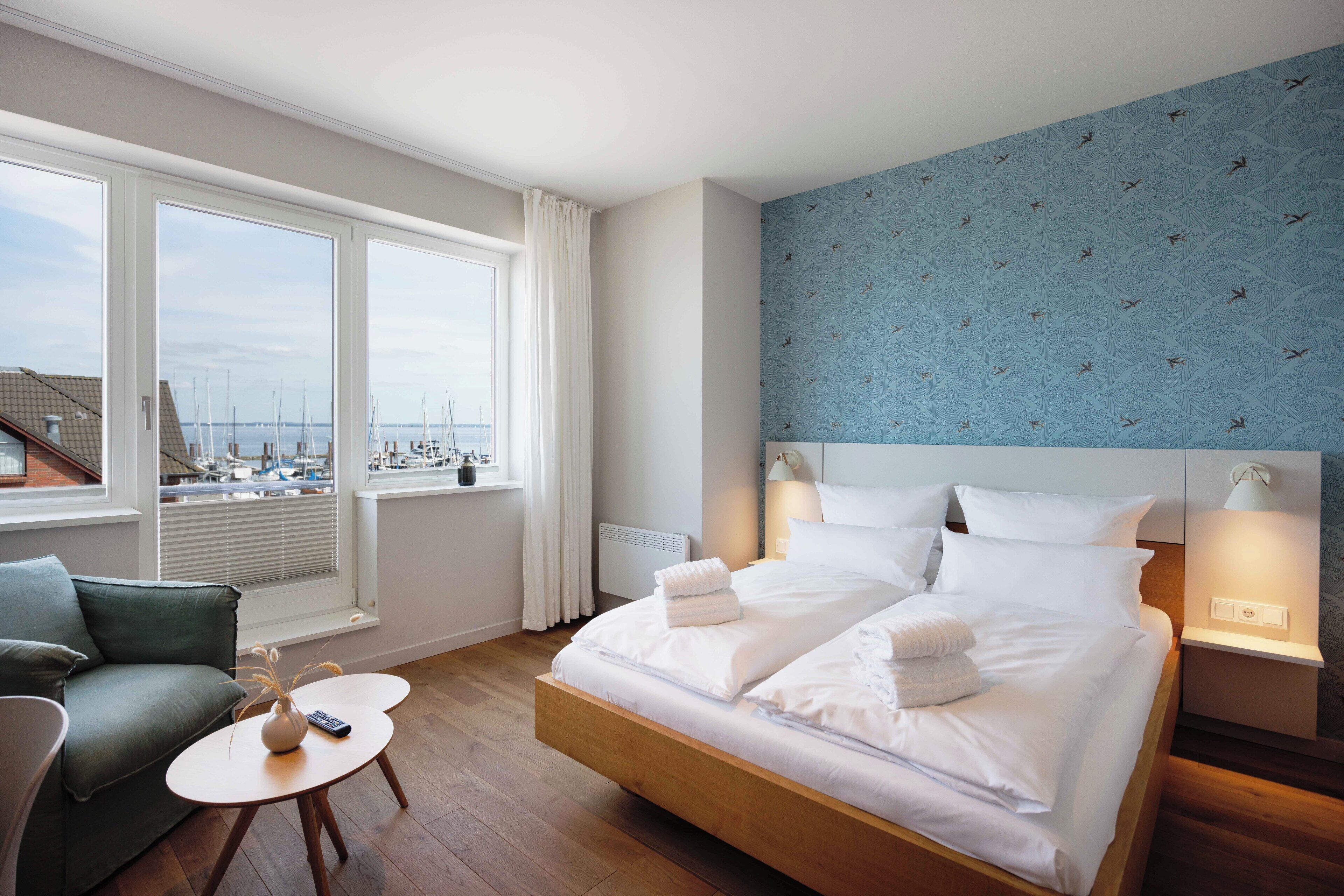A hotel room with a large bed, two bedside tables, a small round table, a sofa, and a view of boats through large windows.