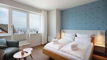 Une chambre d’hôtel avec un grand lit, deux tables de chevet, une petite table ronde, un canapé et une vue sur les bateaux à travers de grandes fenêtres.