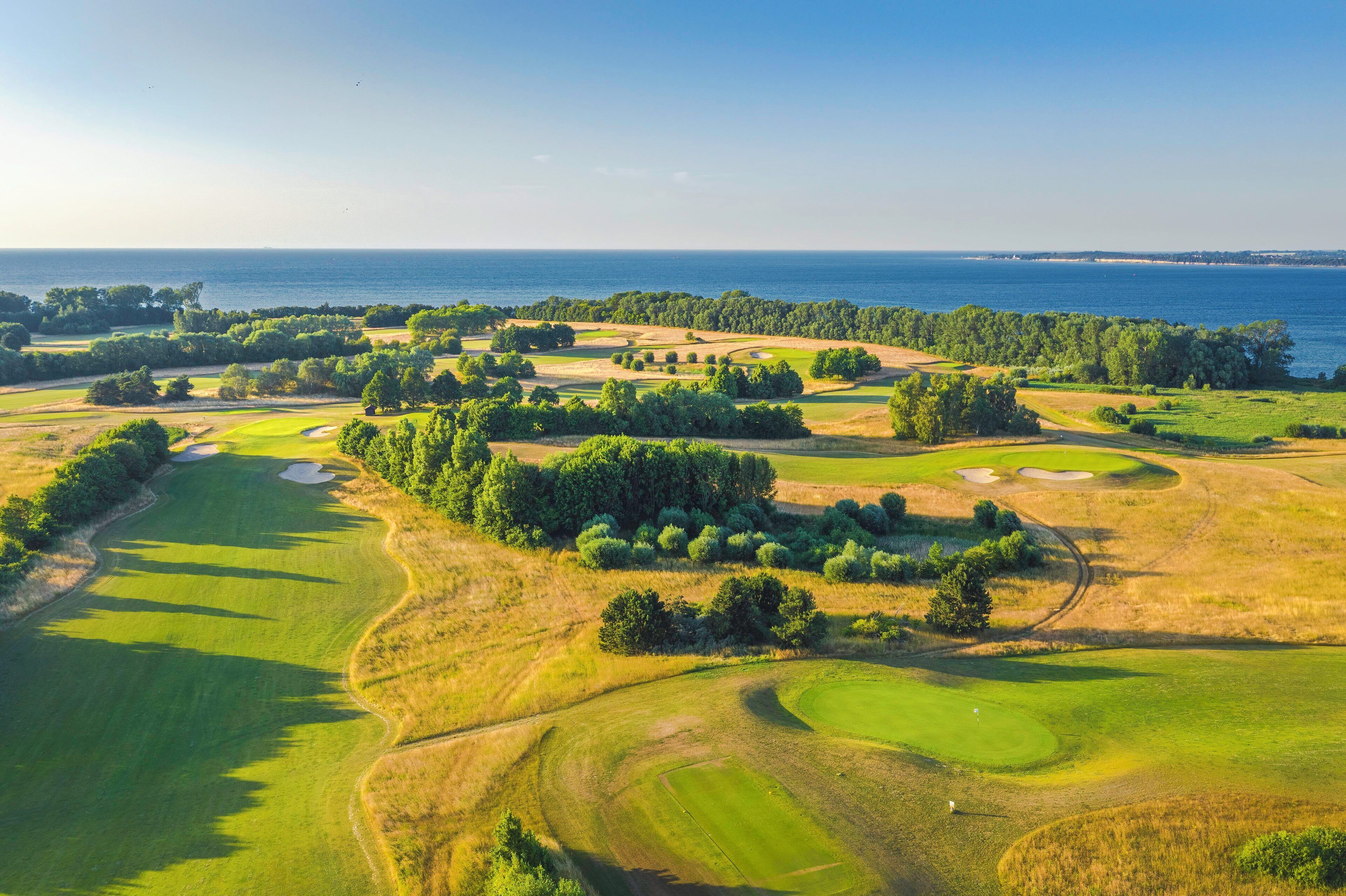 An aerial view of a golf course with multiple greens, sand bunkers, and a large body of water in the background.
