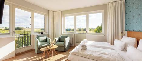 A hotel room with a large bed, two armchairs, a small table, and a view of the countryside through large windows.