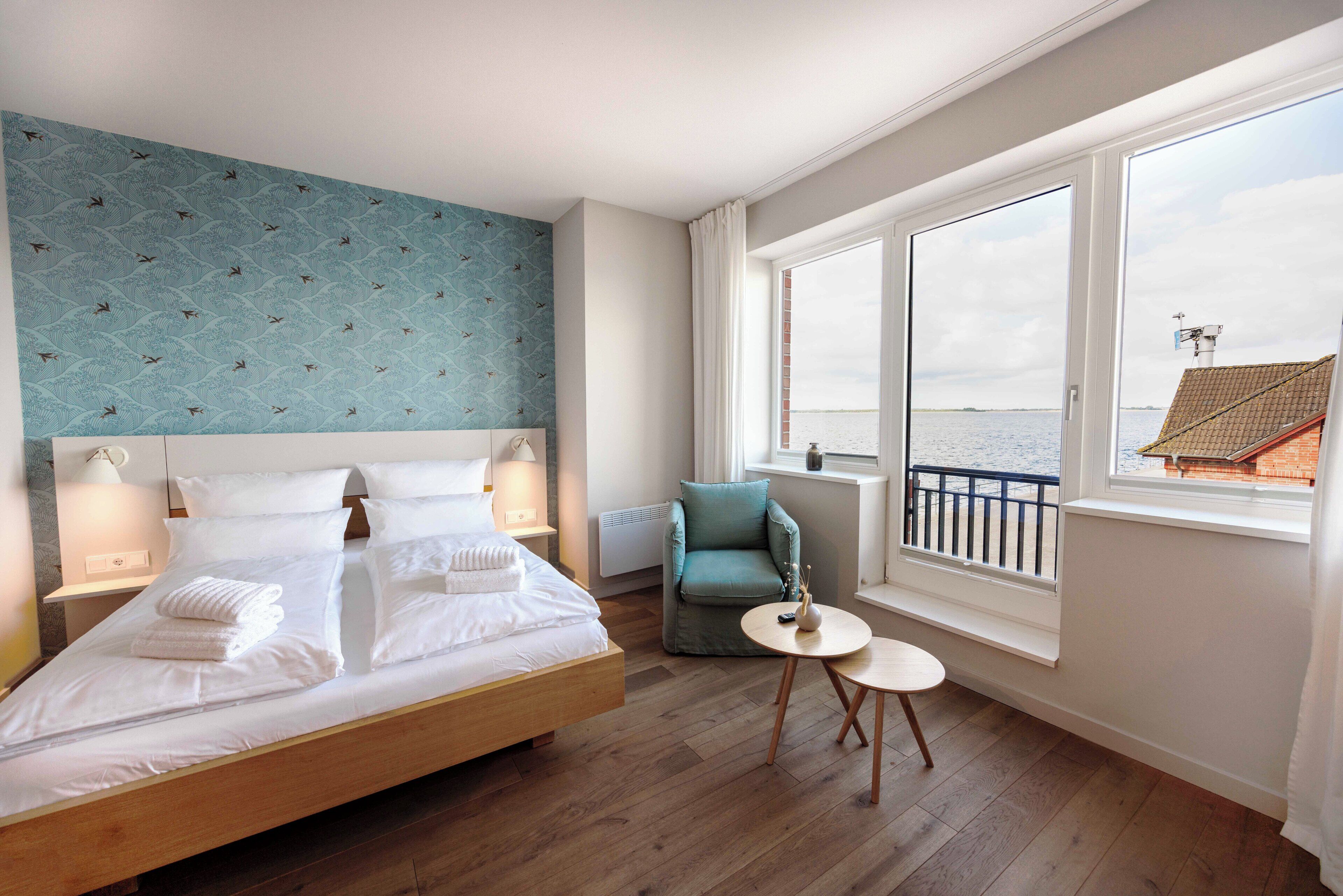 A hotel room with a large bed, a small round table, a chair, and a view of the sea through large windows.