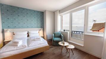 A hotel room with a large bed, a small round table, a chair, and a view of the sea through large windows.