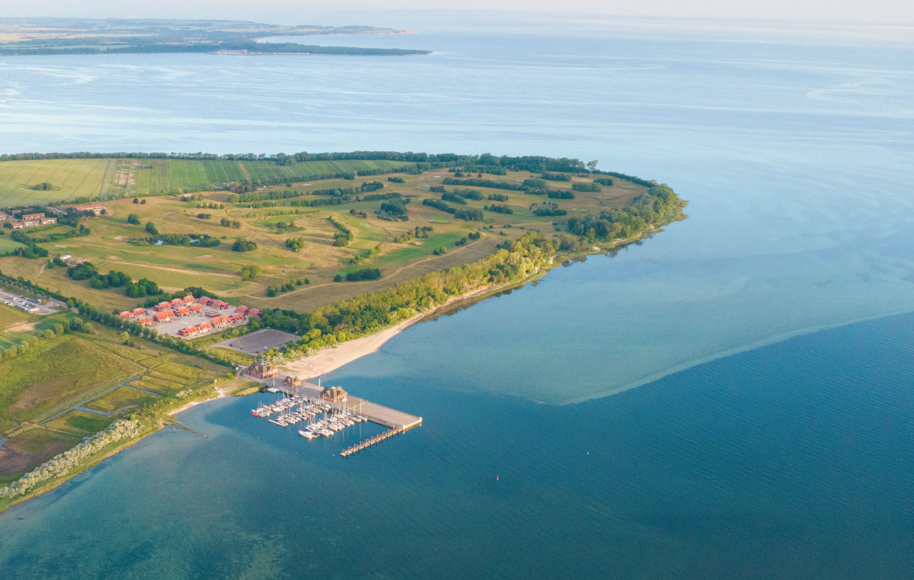 Vue aérienne d’une zone côtière comprenant un port de plaisance, des champs verdoyants et un petit village avec des bâtiments aux toits rouges.