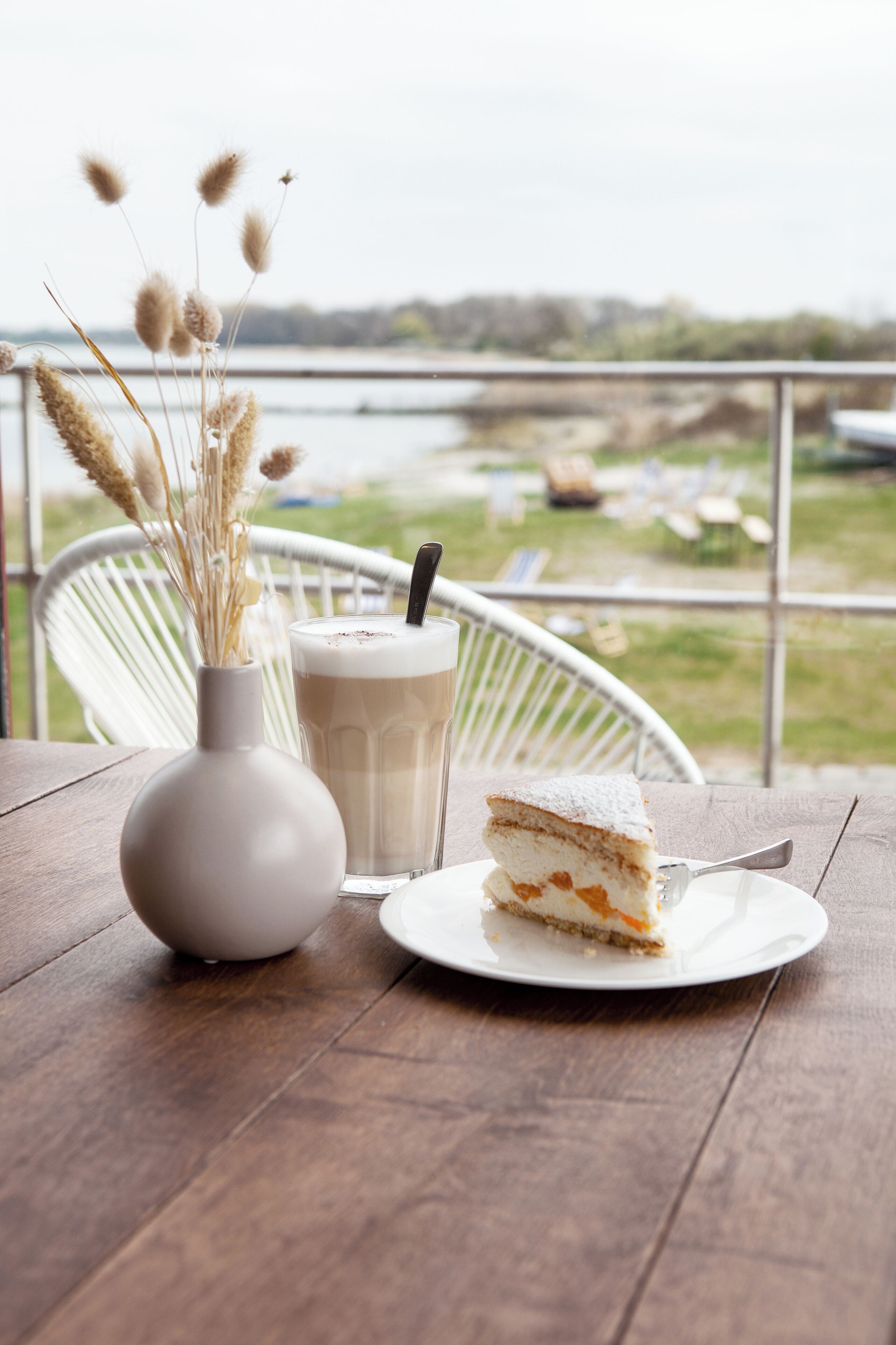 A slice of cake on a plate with a drink and a vase of dried flowers on a wooden table.