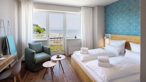 A hotel room with a large window, a bed, a chair, a small table, and a desk.