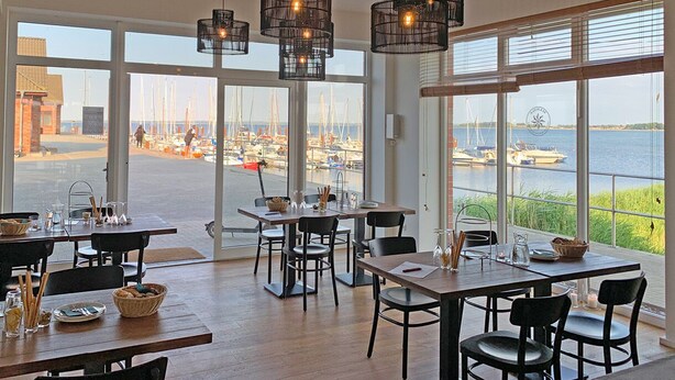 A dining area with large windows overlooking a marina, featuring wooden tables and chairs, and hanging pendant lights.