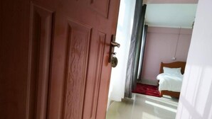 A hotel room with a wooden door, a bed with white bedding, and a red carpet. - The Trumpet Inn (Nyeri)