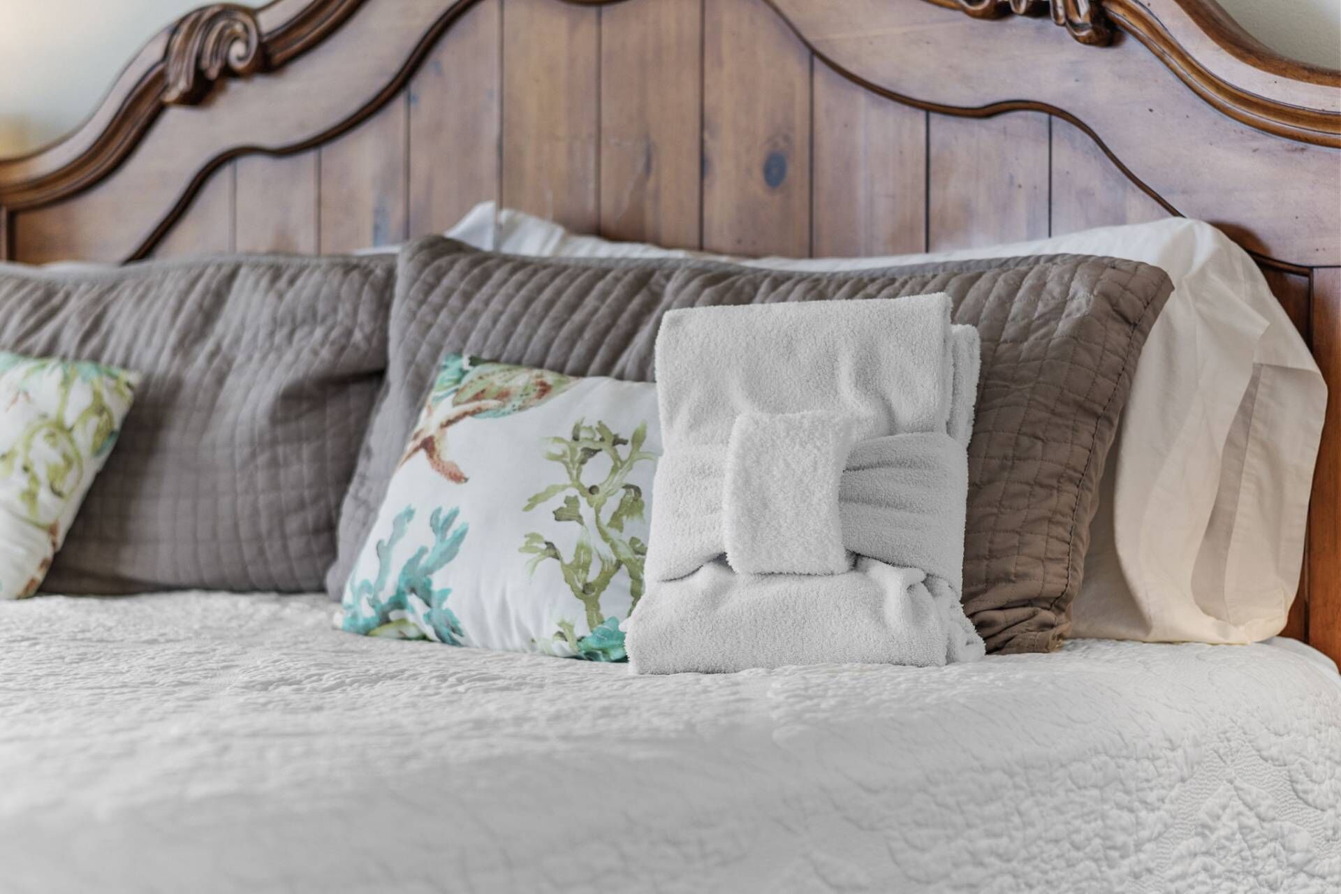 A neatly made bed with a quilted white cover, a folded white towel, and several pillows with floral patterns.
