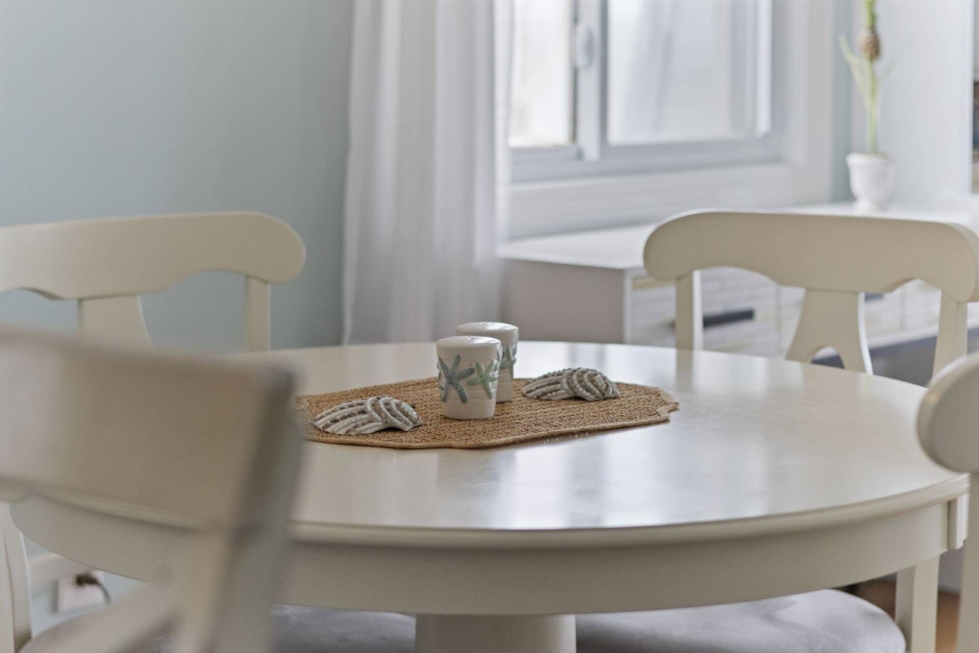 Een ronde witte tafel met een geweven onderzetter, twee witte keramische bekers met sterrendesigns, en twee witte keramieken borden met zeeschelpen.