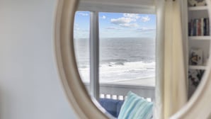 Visão pela janela para o oceano, uma estante e um almofão.