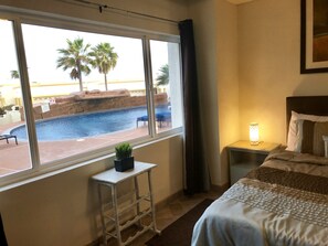 Ein Hotelzimmer mit großem Fenster mit Blick auf einen Swimmingpool und Palmen. Es gibt ein Bett mit Kopfteil, einen Nachttisch mit Lampe und einen kleinen Hocker mit Pflanze.