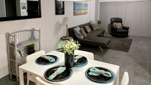 Een eetgedeelte met een witte tafel voor vier personen, een zwarte hanglamp en een bank op de achtergrond.