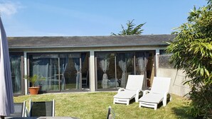 Ein modernes Haus mit einer verglasten Terrasse, zwei weißen Lounge-Stühlen, einem Blumentopf und einem großen Sonnenschirm.