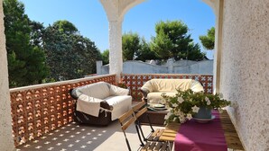 A balcony with a wicker sofa, a small table with a vase of flowers, and a view of trees and a building. - Villa AURORA, beautiful sea view! 400mt from the sandy beach. (Lizzano)