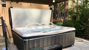 A hot tub with a built-in seat and steps, surrounded by a wooden deck and a forested area. - CdA Hotspot - Hot Tub, Pool, & Sauna (Coeur d Alene)