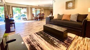 A modern living room with a black sofa, a coffee table, and a TV stand. There is a dining area with a table and chairs visible through the glass doors. - CdA Hotspot - Hot Tub, Pool, & Sauna (Coeur d Alene)