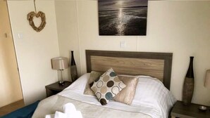 Un lit bien fait, agrémenté d’une décoration en forme de cœur, d’un tableau représentant un coucher de soleil et d’un vase posé sur la table de chevet.