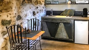 A rustic kitchen with stone walls, a wooden ceiling, and a small dining area with a metal chair and table. - Former "U FRAGNU" wine press (Omessa)