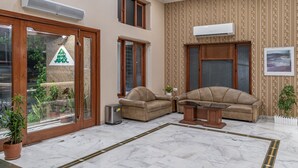 Un salon spacieux avec deux canapés, une table basse et une grande fenêtre. Des unités de climatisation sont fixées aux murs, et une plante en pot est posée sur le sol.