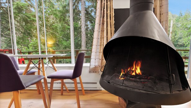 Una chimenea moderna con fuego, suelo de madera y vistas a árboles a través de amplios ventanales.