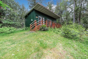A green cabin with a wooden staircase leading to a door, surrounded by dense forest.