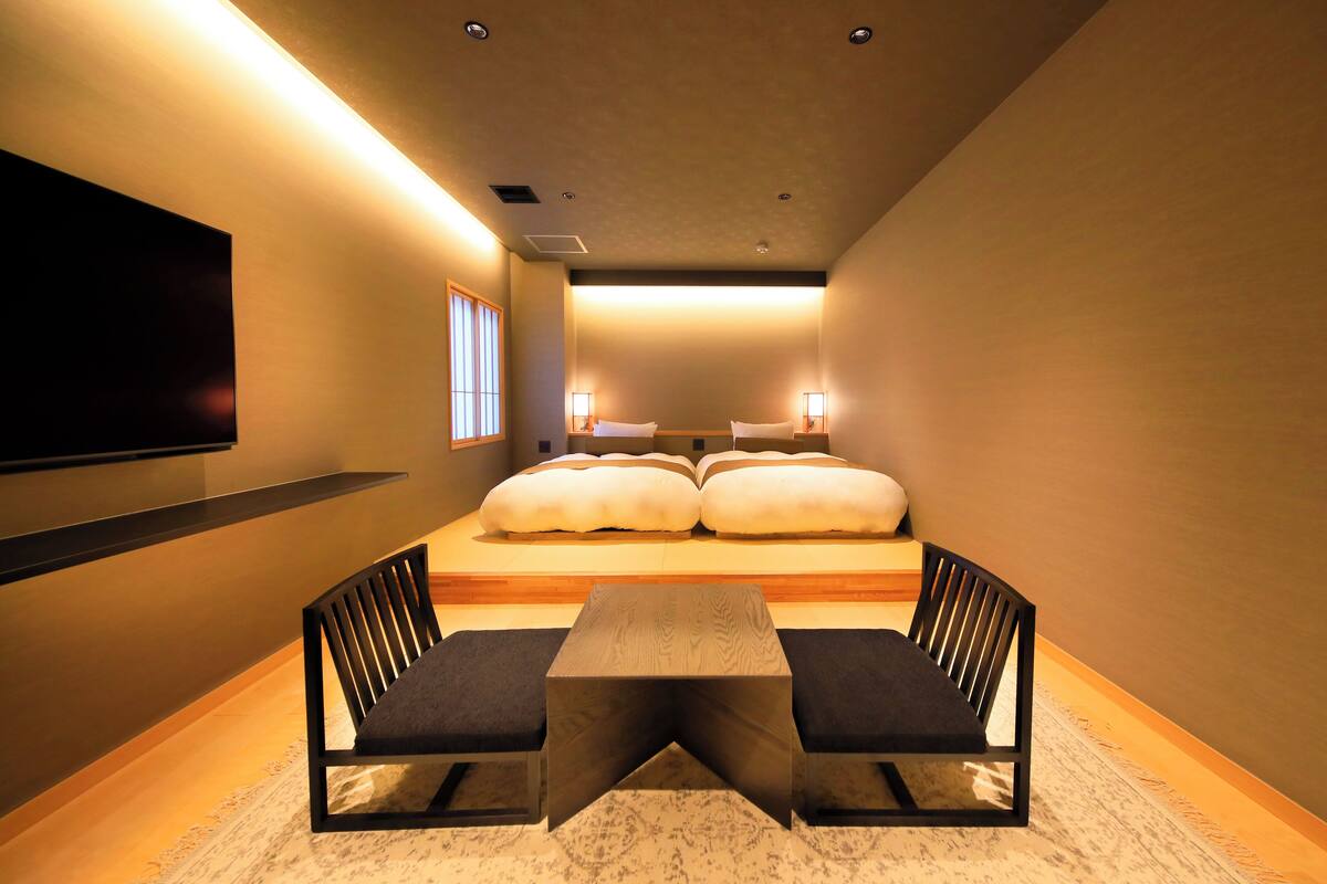 A modern hotel room with a large bed, two bedside tables, a flat-screen TV mounted on the wall, and a small seating area with two chairs.