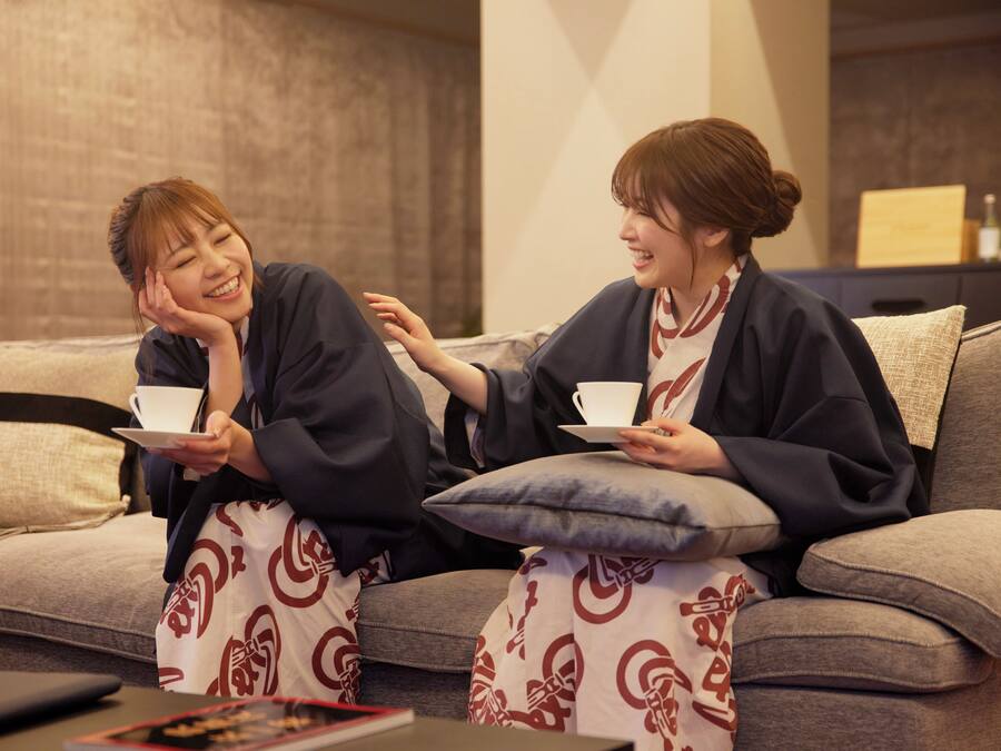 Two people sitting on a sofa with cups, in a room with a brick wall and a wooden cabinet in the background.
