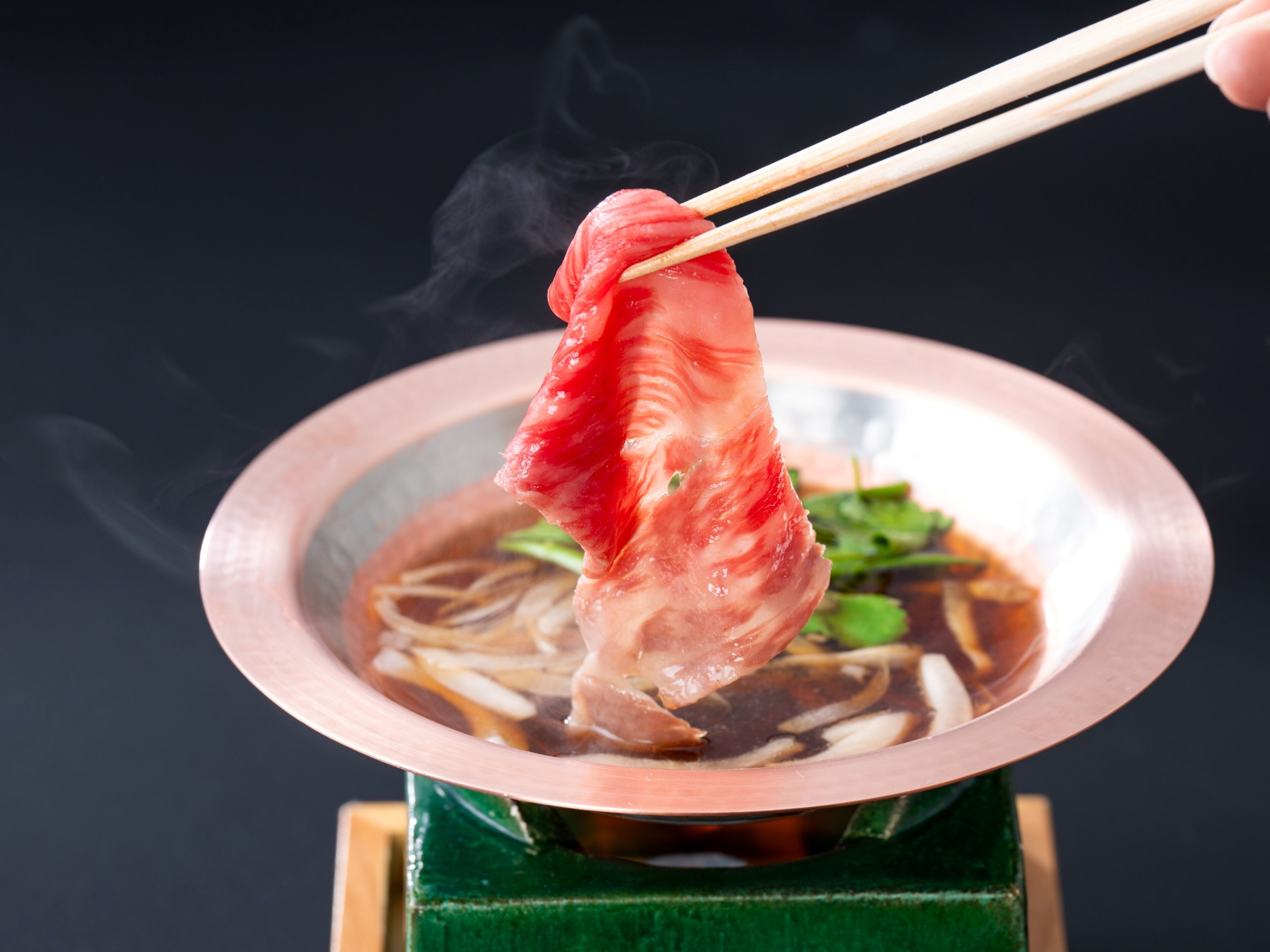 Un tazón de sopa con palillos levantando un trozo de carne, vapor elevándose del tazón y una base verde.