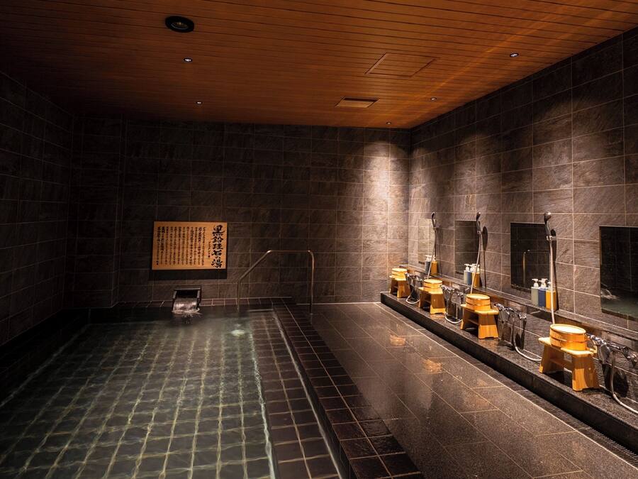 A modern bathroom with dark tiled walls, a wooden ceiling, and a long bench with sinks and mirrors.