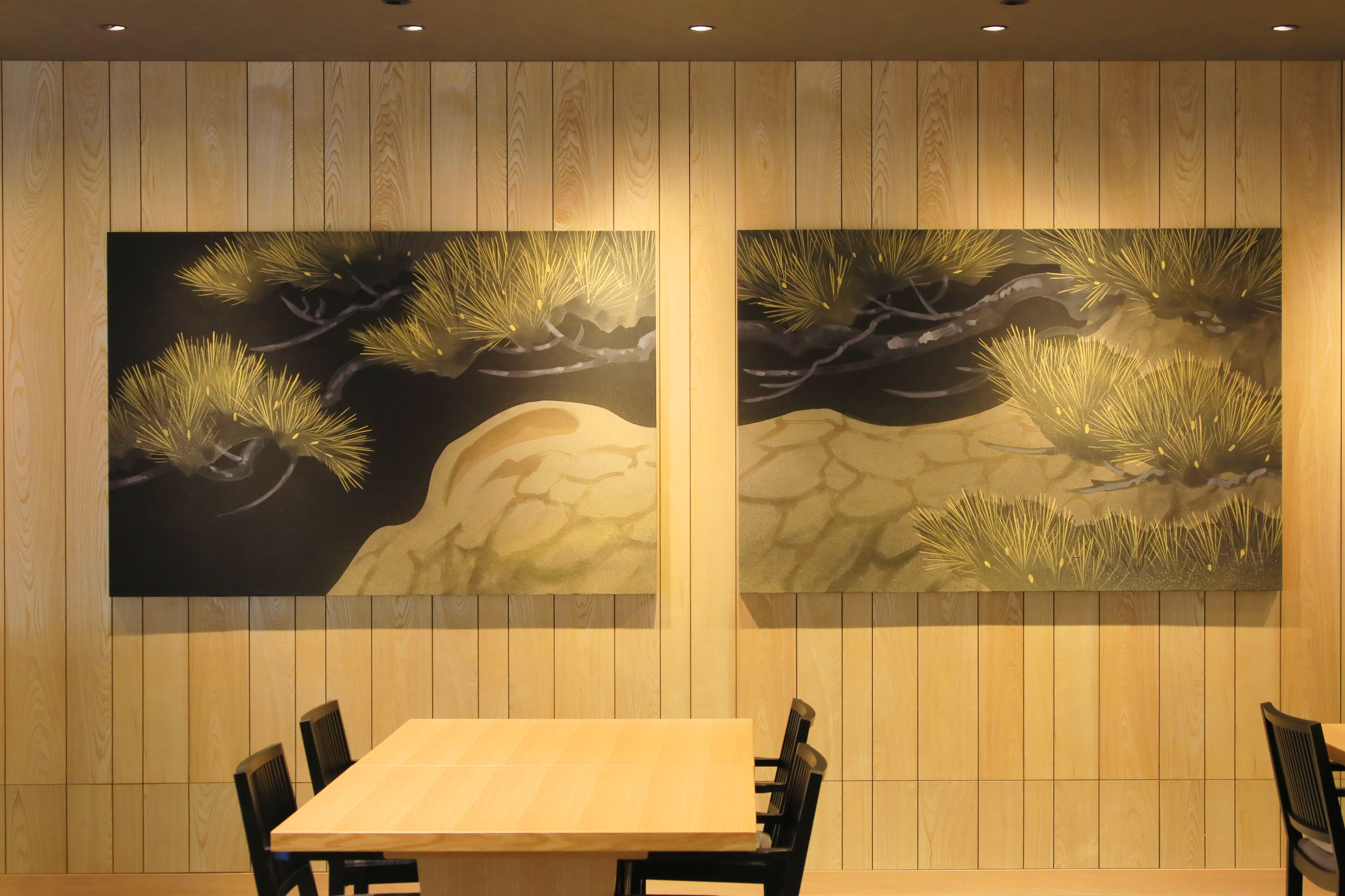 A dining area with a wooden table and chairs, and two large wall-mounted paintings of pine trees.