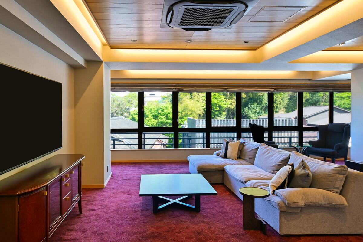 A living room with a sectional sofa, a coffee table, a TV, and a large window with a view of greenery.