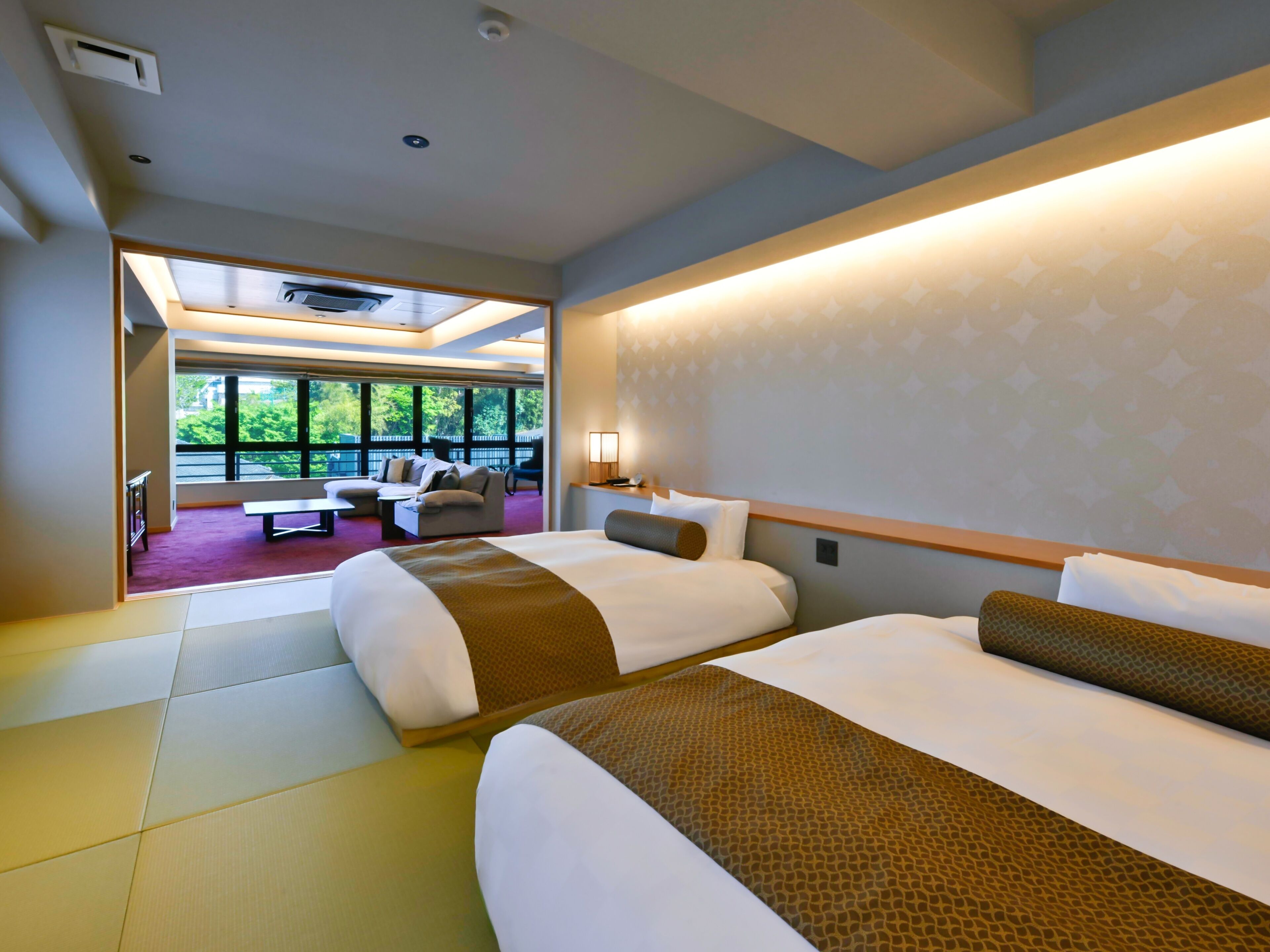 A hotel room with two beds, a sofa, a dining table, and large windows offering a view.