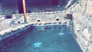 Une piscine bordée de pierres, avec un banc en bois et un mur de pierres percé de petites ouvertures.