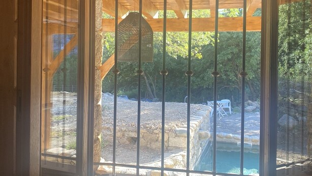 Une vue à travers une fenêtre ornée de barreaux de métal, dévoilant un espace extérieur avec une structure en pierre, une piscine et de la verdure.