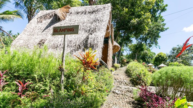 A thatched-roof structure labeled "GLAMPING" with a stone pathway leading to it, surrounded by lush greenery and palm trees. - Taman Asta Gangga by ecommerceloka (Sidemen)