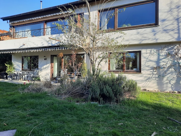 Une maison à deux étages dotée d’un balcon, d’une terrasse avec des meubles d’extérieur et d’un jardin agrémenté de diverses plantes.