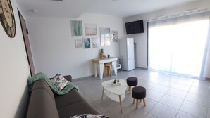 Ett modernt vardagsrum med en brun soffa, ett litet vitt bord och två svarta pallar. På väggen sitter en platt-tv, och i rummet finns ett kylskåp och ett fönster med tunna, genomskinliga gardiner.