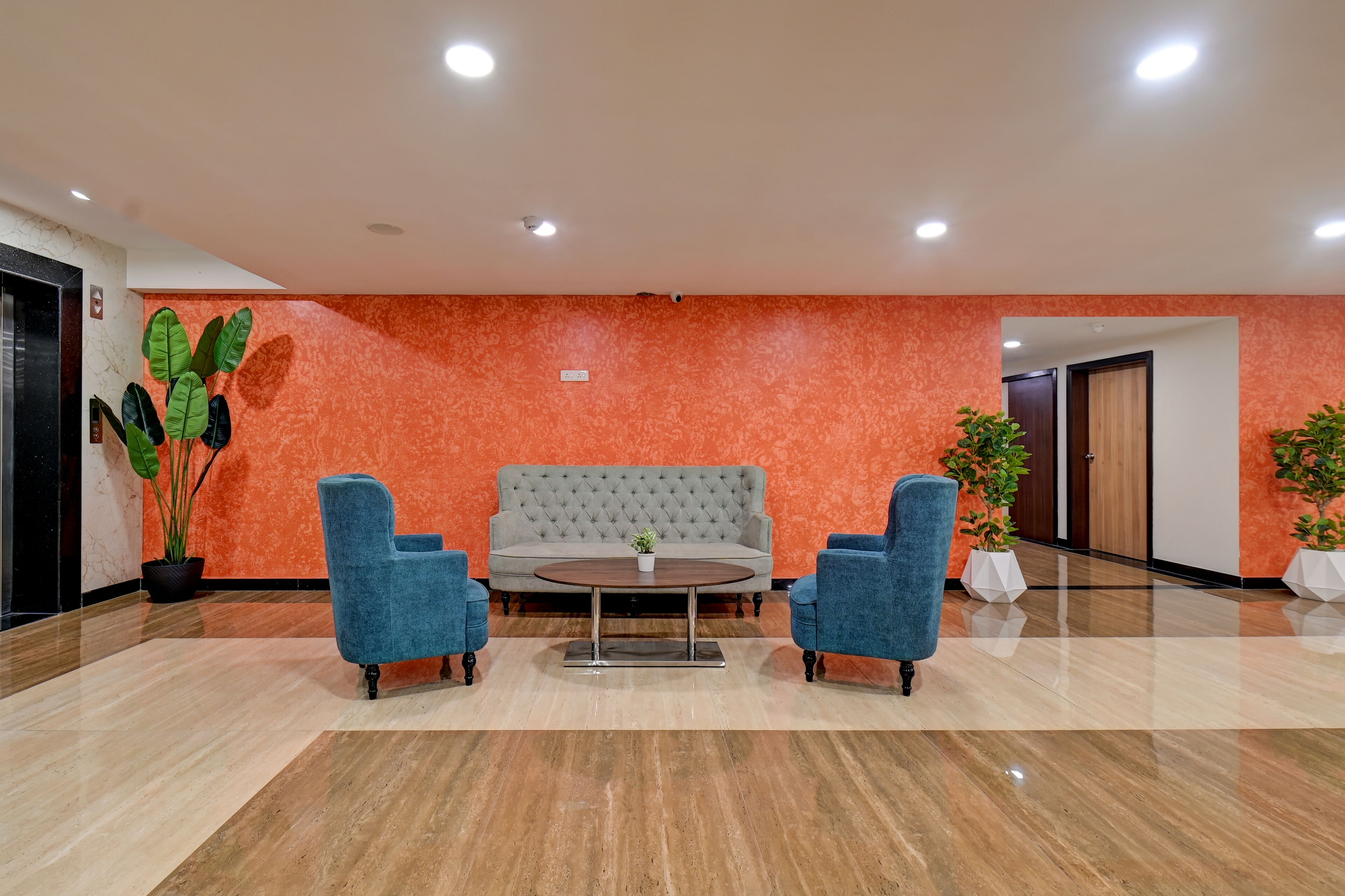 A lobby with a grey tufted sofa, two blue armchairs, a round coffee table, and potted plants.